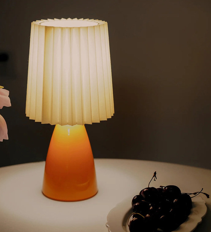 Small ceramic table lamp in burnt orange with a pleated beige shade, lit on a dark table