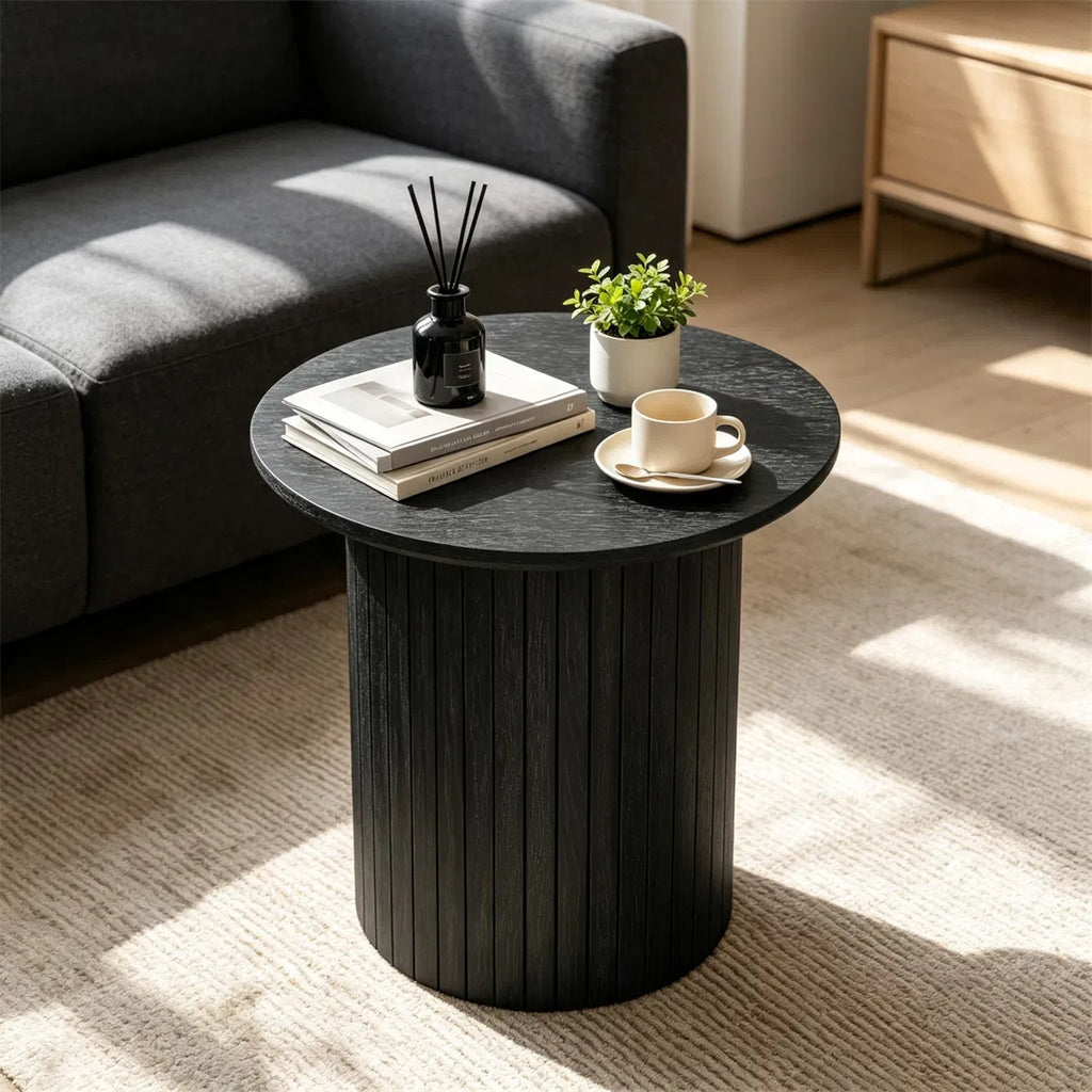 Black fluted pedestal side table by a grey sofa, topped with books, a reed diffuser and a cup