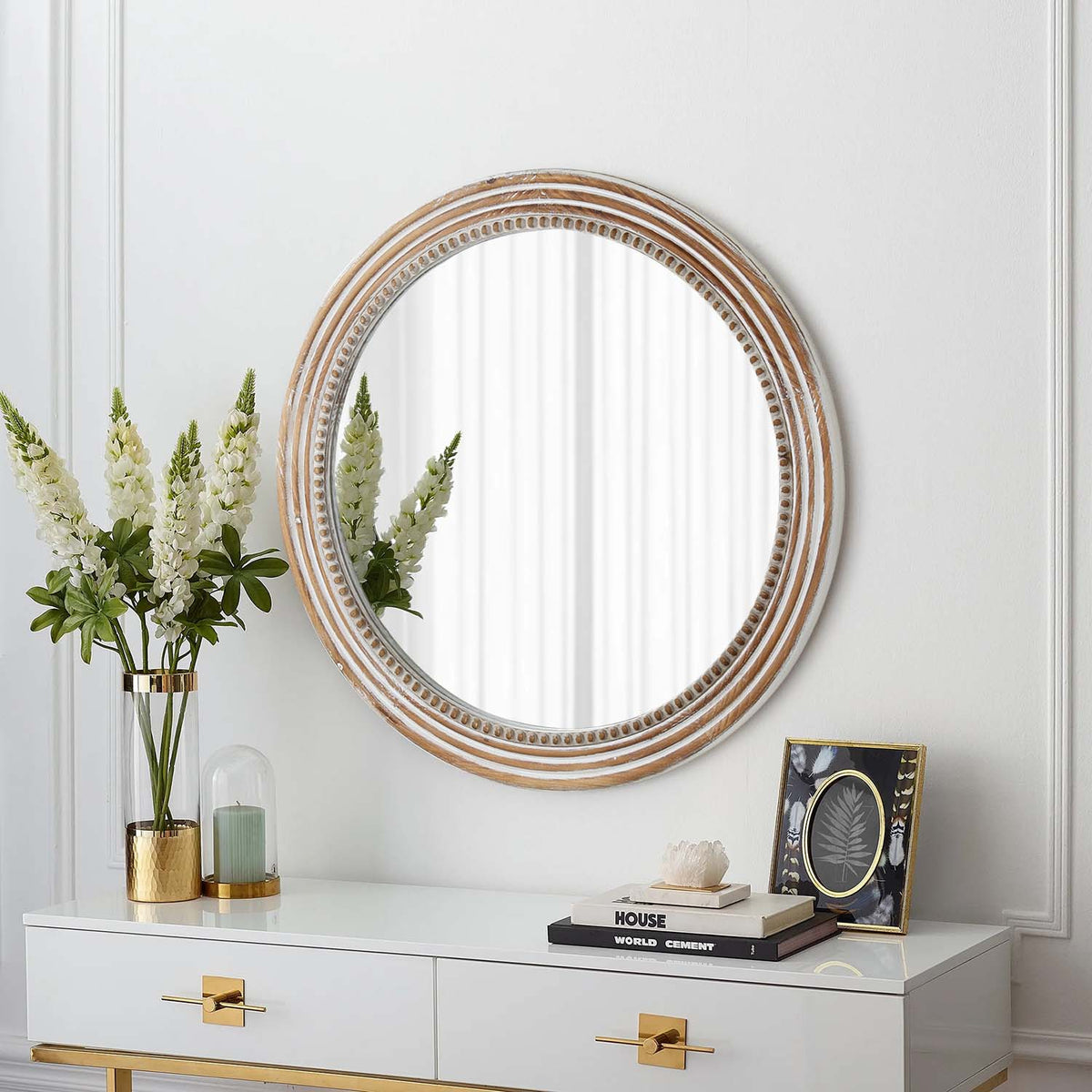 Whitewashed beaded round mirror above a white console with white flowers in a gold vase
