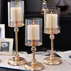 Three gold pedestal hurricane candle holders in graduated heights, lit on a table runner beside framed prints