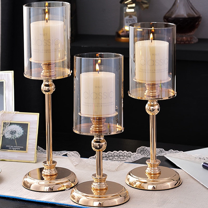 Three gold pedestal hurricane candle holders in graduated heights, lit on a table runner beside framed prints