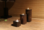Three charred paulownia tealight holders lit on a wooden shelf before a bamboo blind