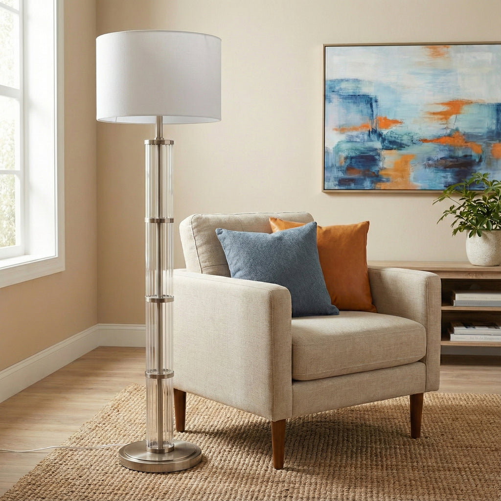 Glass column floor lamp with white drum shade beside an armchair with blue and rust pillows