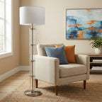 Glass column floor lamp with white drum shade beside an armchair with blue and rust pillows