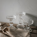 Three clear glass pillar candle holders at three heights on a linen cloth against a grey wall