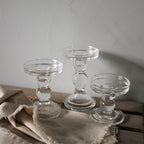 Three clear glass pillar candle holders at three heights on a linen cloth against a grey wall