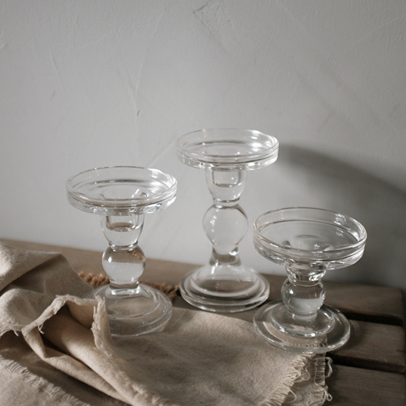 Three clear glass pillar candle holders at three heights on a linen cloth against a grey wall