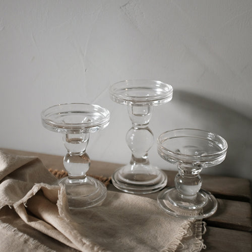 Three clear glass pillar candle holders at three heights on a linen cloth against a grey wall