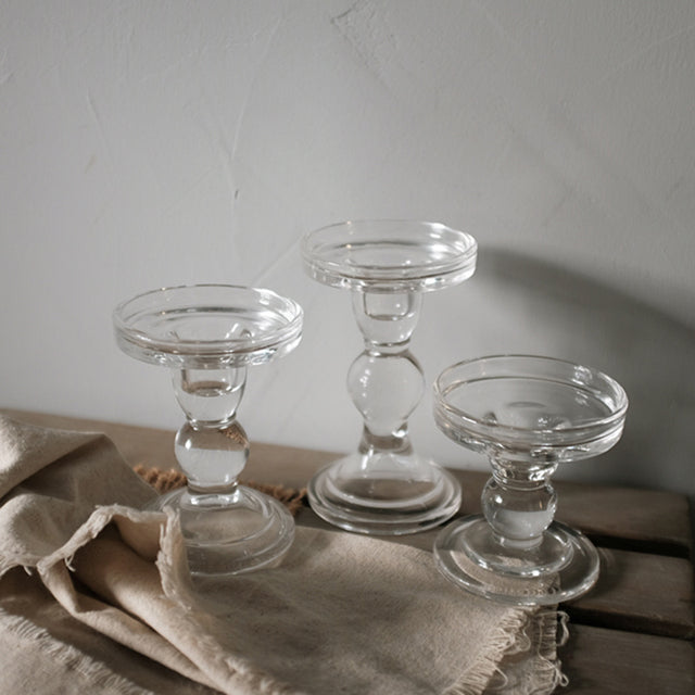 Three clear glass pillar candle holders at three heights on a linen cloth against a grey wall