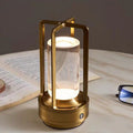 Brass cordless lantern table lamp with clear cylinder, lit on a marble table beside an open book