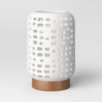 Large cutout ceramic lantern candle holder in speckled white on a brown foot, on a plain light grey background