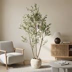 Faux olive tree in a stone planter, not included, beside a linen armchair and table lamp