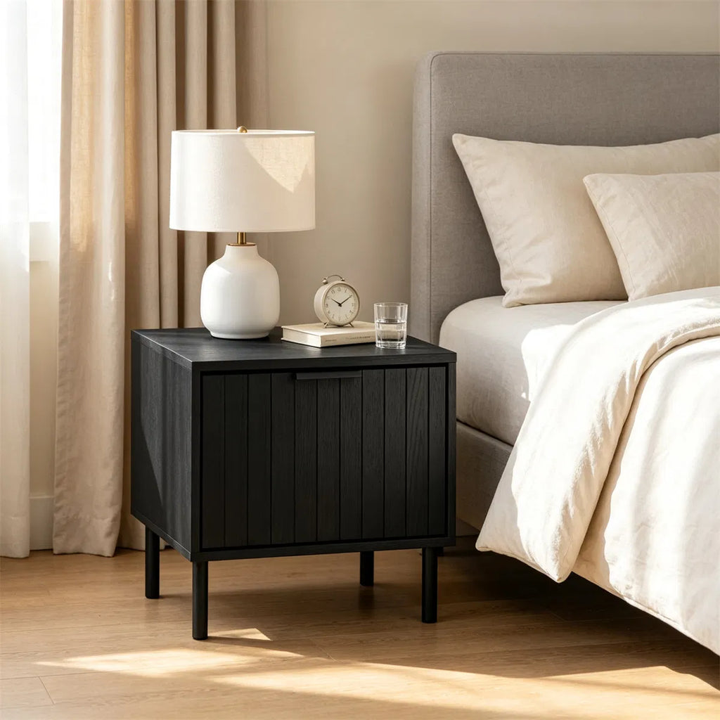 Fluted black nightstand beside a bed with a white ceramic lamp and clock