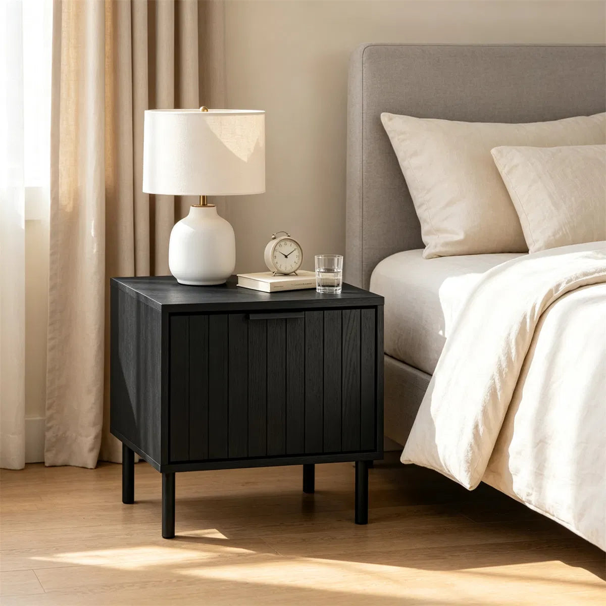 Fluted black nightstand beside a bed with a white ceramic lamp and clock