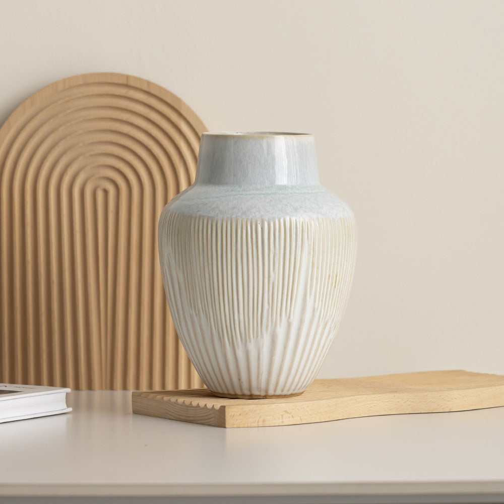 Fluted ceramic vase with pale blue shoulder on a wavy wooden board in front of a ribbed wooden arch