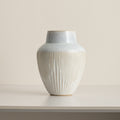 Fluted ceramic vase with pale blue glazed shoulder and cream ribbed body on a white table against a beige wall