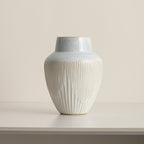 Fluted ceramic vase with pale blue glazed shoulder and cream ribbed body on a white table against a beige wall