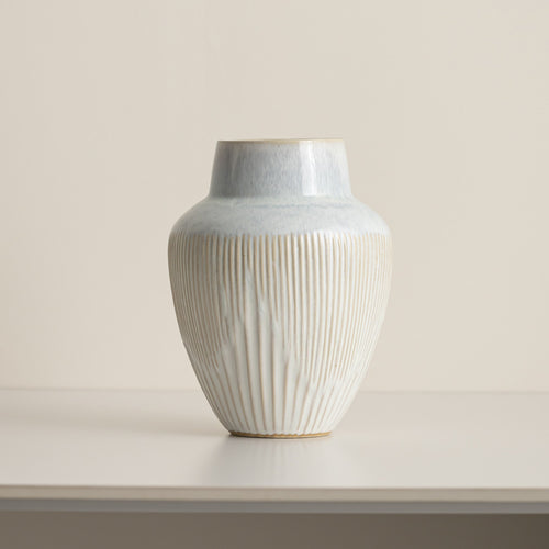 Fluted ceramic vase with pale blue glazed shoulder and cream ribbed body on a white table against a beige wall