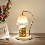 Candle warmer lamp lit over a gold jar candle beside pink roses and a white picture frame