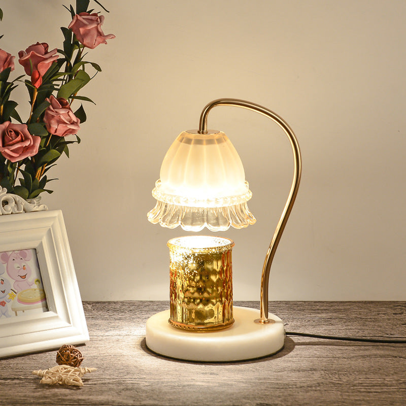 Candle warmer lamp lit over a gold jar candle beside pink roses and a white picture frame