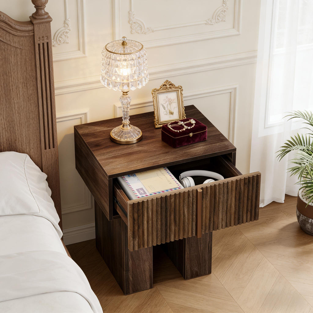 Fluted pedestal nightstand with the drawer open beside a bed, holding a notebook and headphones