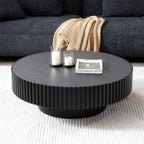 Matte black fluted drum coffee table with candles and a magazine before a charcoal sofa and tan throw