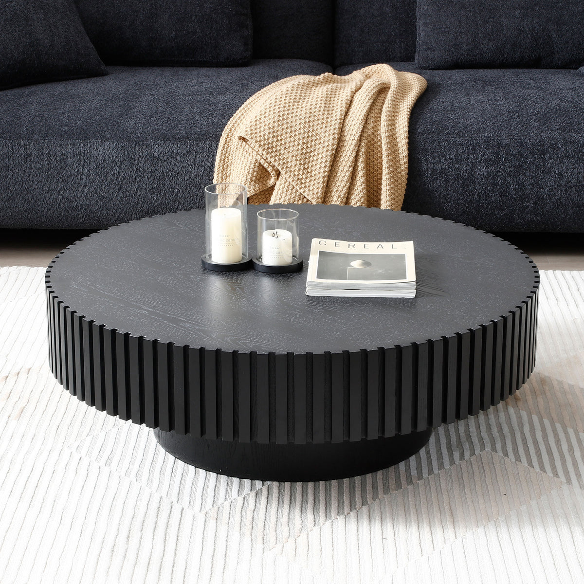 Matte black fluted drum coffee table with candles and a magazine before a charcoal sofa and tan throw