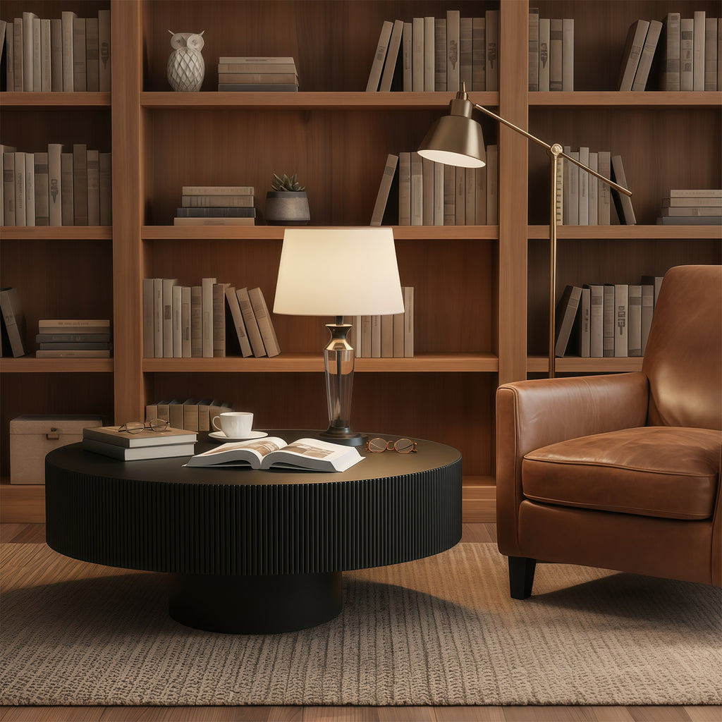 Black fluted coffee table in a book-lined room beside a brown leather armchair and table lamp