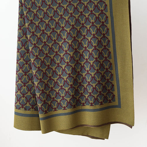 Olive knit throw blanket with interlocking ring pattern and wide border, hanging against a white wall