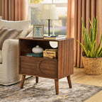 Walnut arched-panel nightstand beside a linen armchair, a lamp and books on top, in a sunlit living room