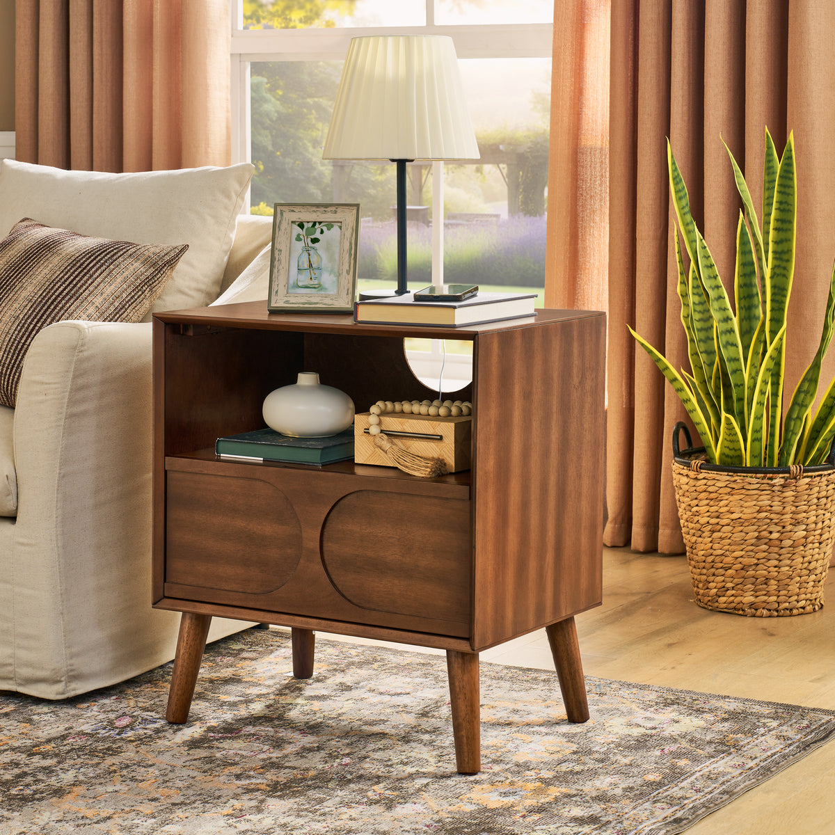 Walnut arched-panel nightstand beside a linen armchair, a lamp and books on top, in a sunlit living room