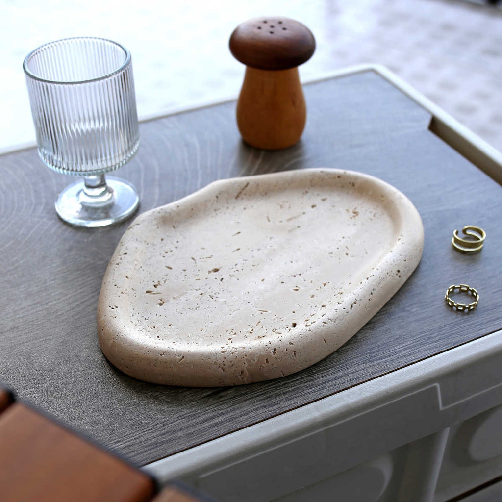 Beige travertine catchall tray with two rings, beside a ribbed glass and a wooden shaker