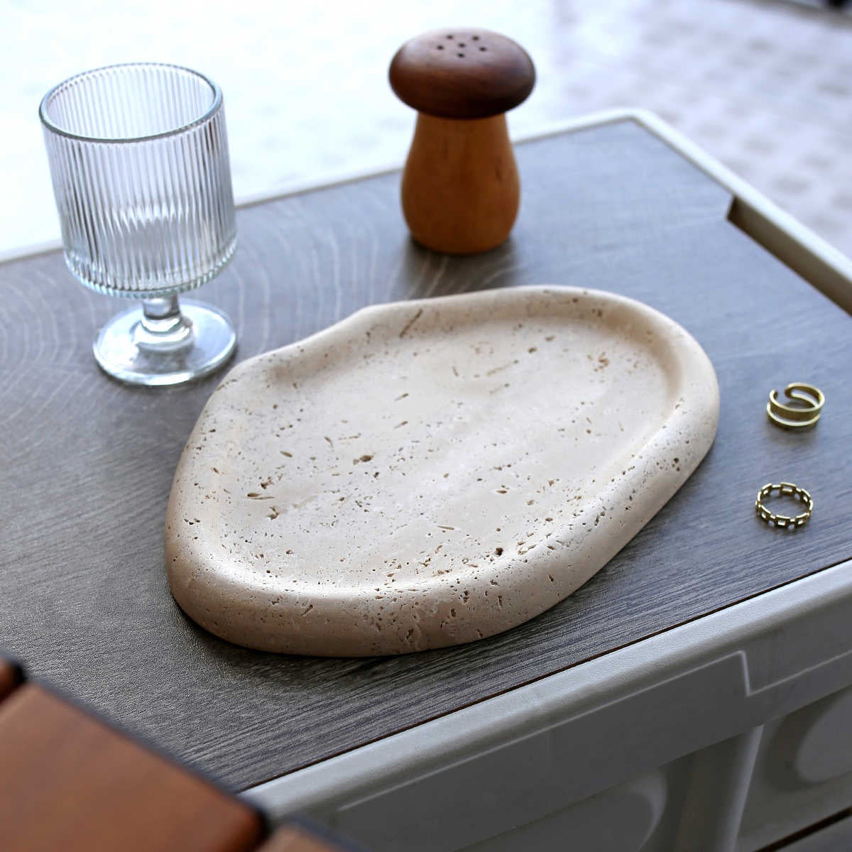 Beige travertine catchall tray with two rings, beside a ribbed glass and a wooden shaker