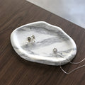 Pebble-shaped stone catchall tray in White marble with gray veins, holding earrings, on a dark wood table