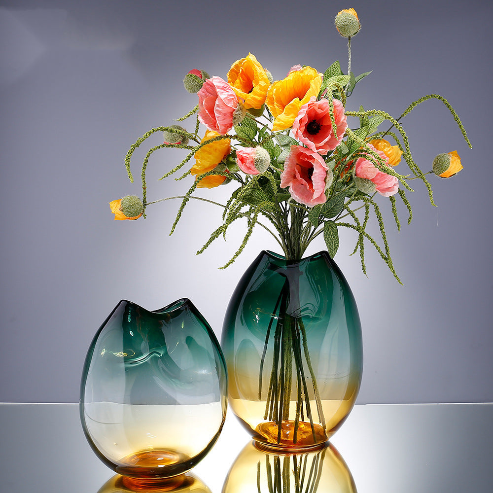 Pinched ombre glass vase in teal and amber beside a taller version holding poppies, on a reflective surface