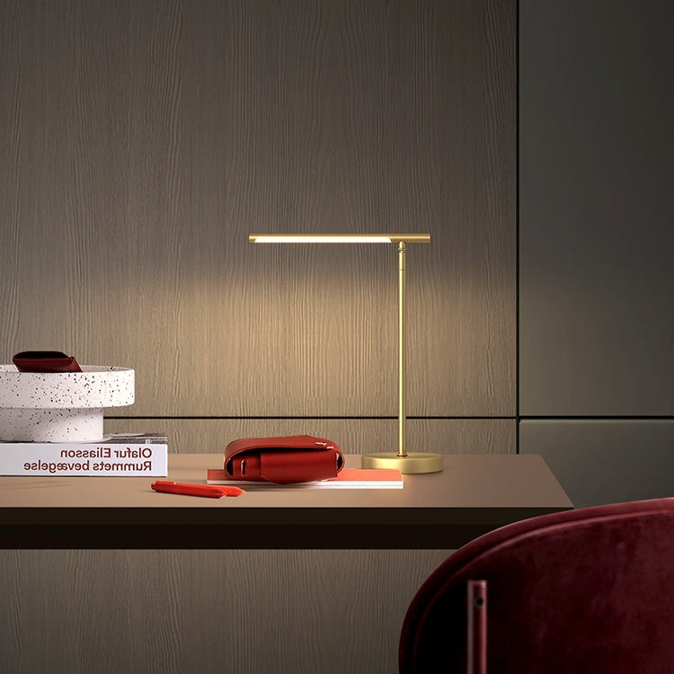Brass light-bar desk lamp lit on a dark desk beside stacked books and a red pouch