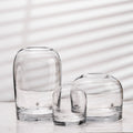 Rounded glass bud vases in clear, three heights side by side on white marble with striped window shadows