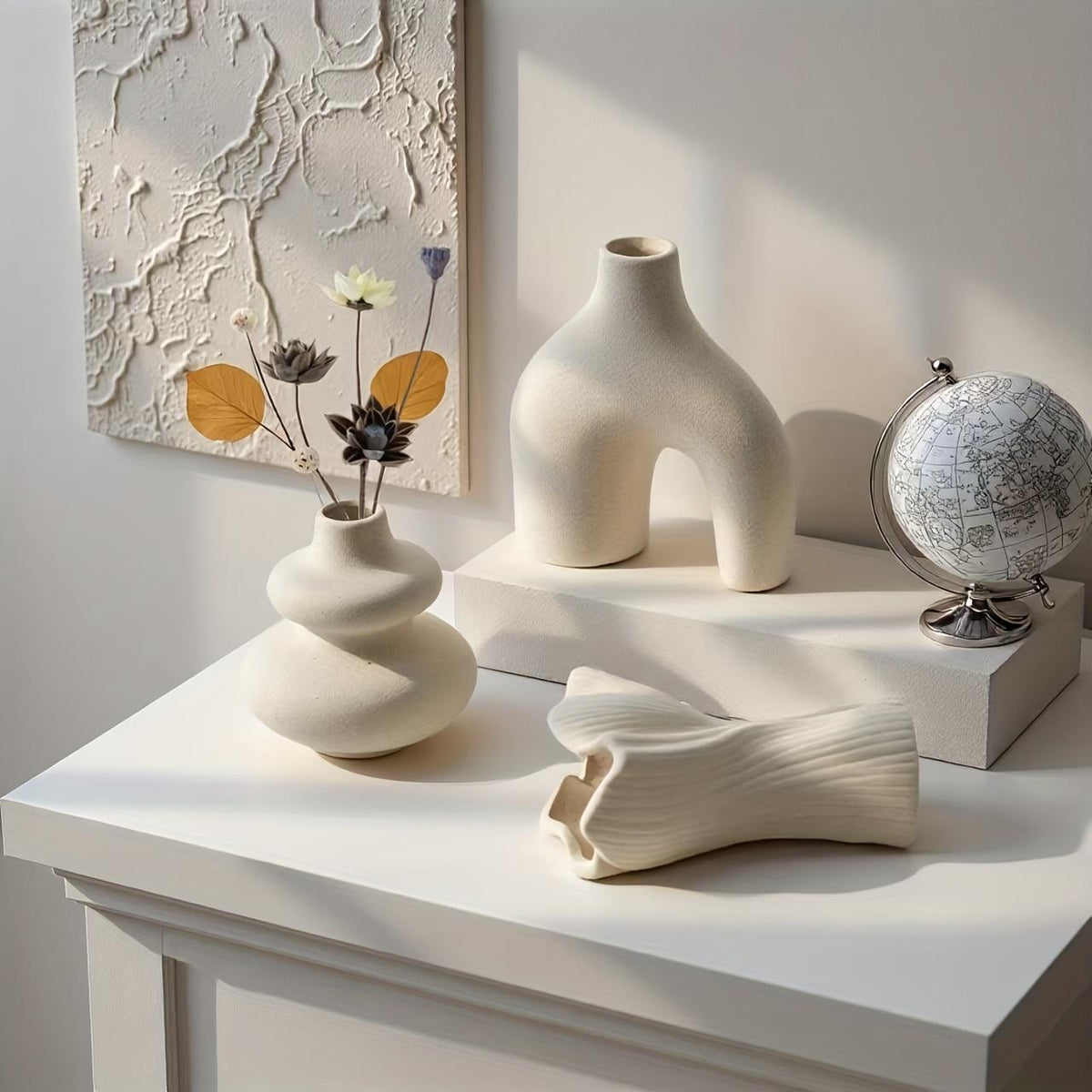 Matte white swirl vase with dried stems, arch vase on a riser and fan vase on its side on a white console