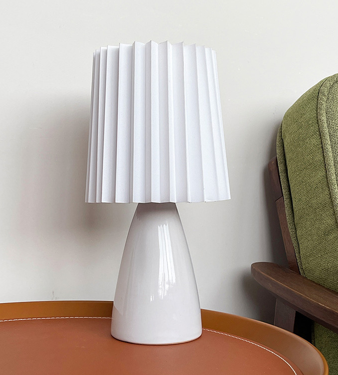 Small white ceramic table lamp with a white pleated shade on a round leather-top table