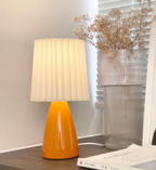 Burnt orange pleated lamp lit by a window beside a ribbed glass vase