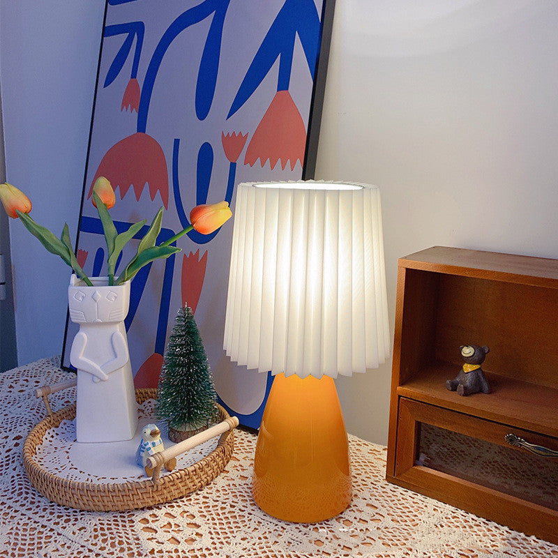 Burnt orange ceramic lamp with pleated shade lit on a lace cloth beside tulips
