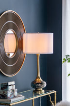 Smoked glass gourd table lamp lit on a glass console beside a round ribbed mirror