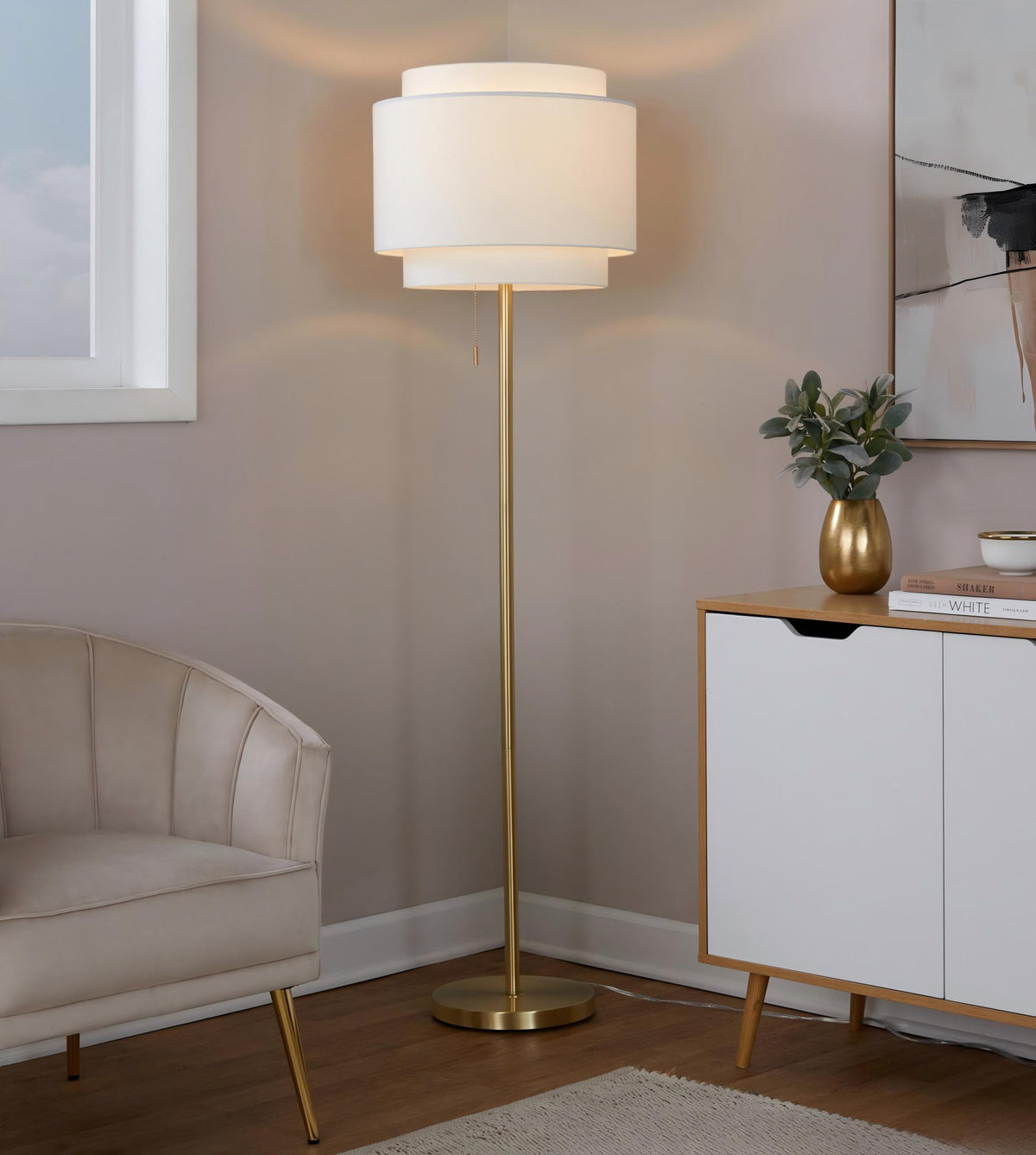 Tiered linen shade floor lamp lit in a living room corner beside a velvet armchair and white sideboard