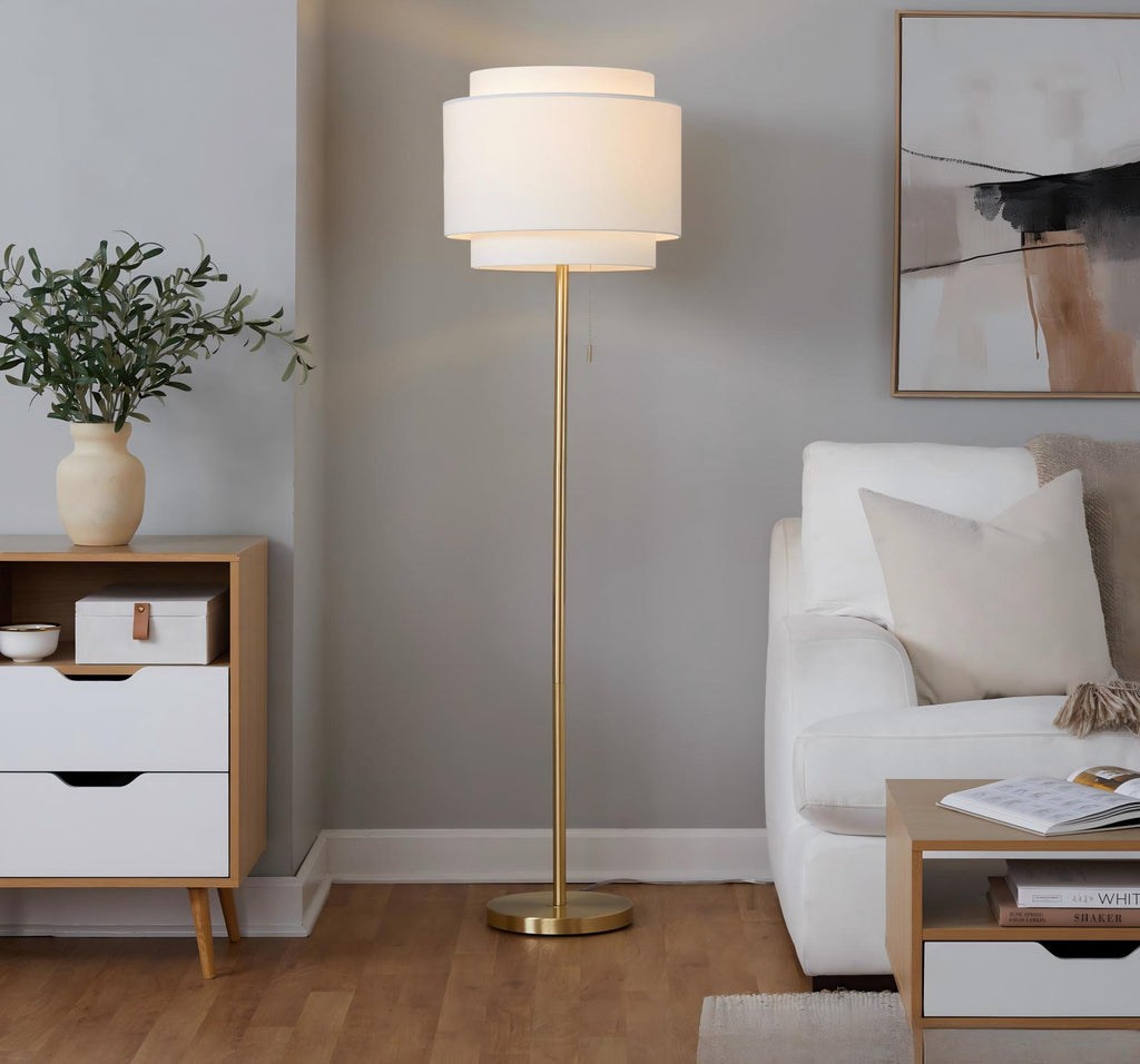 Gold floor lamp with off-white two-tier shade lit beside a white sofa and wood storage cabinet