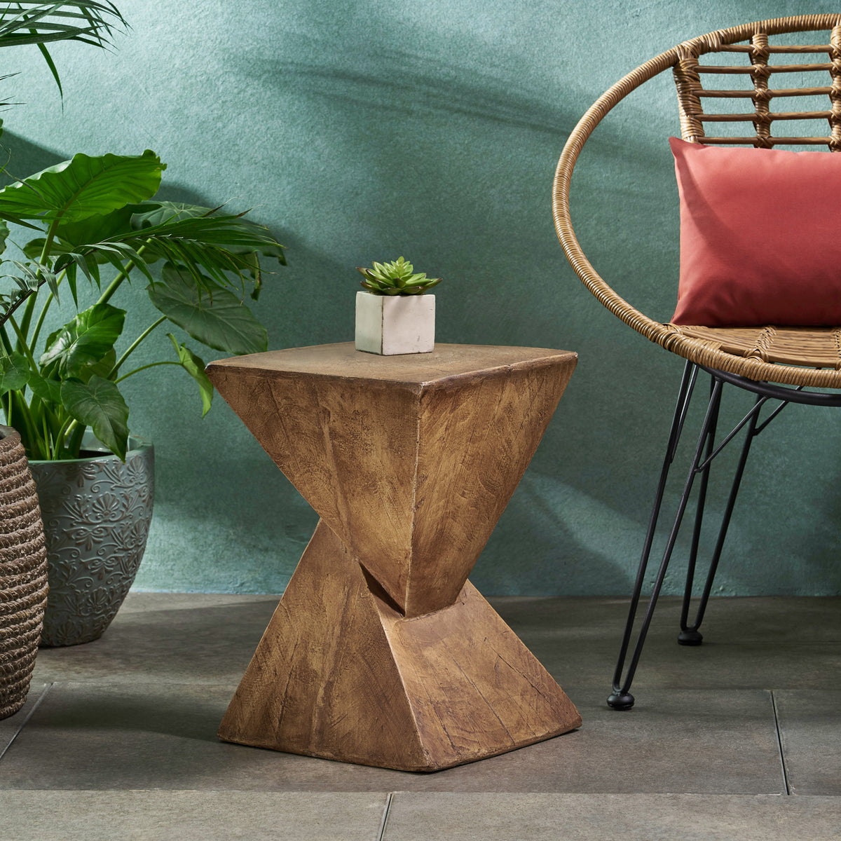 Wood-look twisted side table against a green wall beside a rattan chair and a potted plant