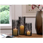 Black rod candle holders lit on a table beside a smoked glass vase