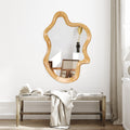 Wavy irregular wall mirror with natural wood-finish frame on a white wall above a woven bench