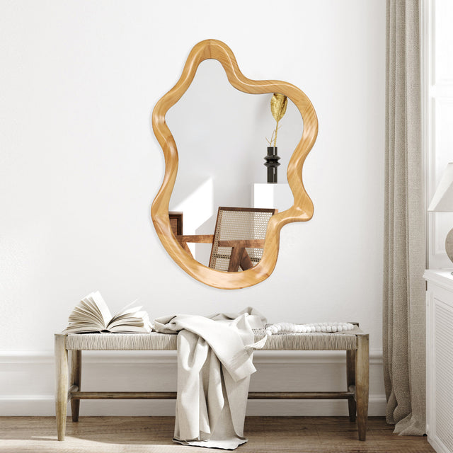 Wavy irregular wall mirror with natural wood-finish frame on a white wall above a woven bench