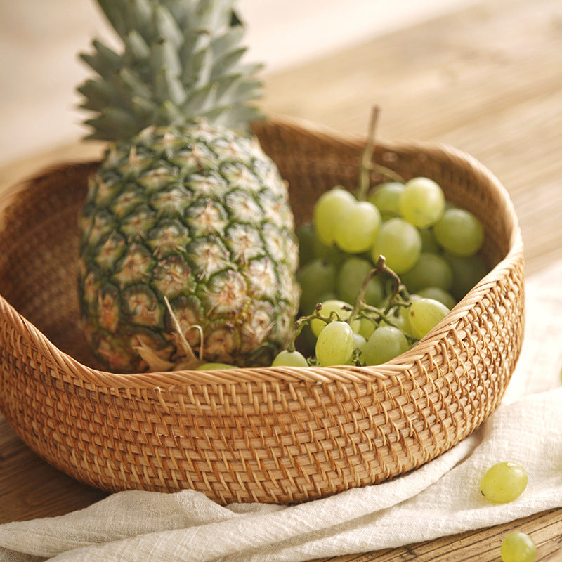 Round woven fruit basket holding a pineapple and green grapes on a wooden table
