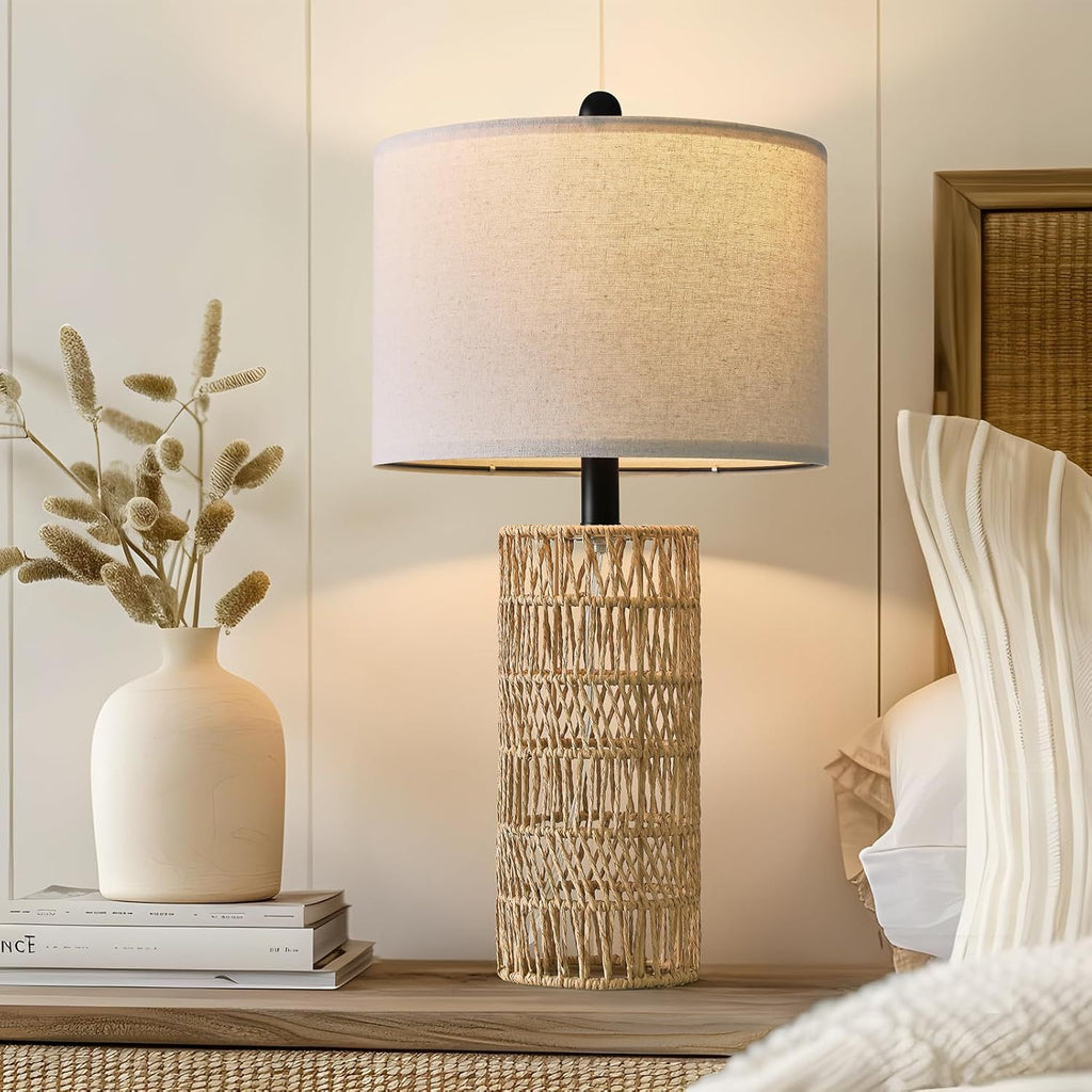 Woven rattan table lamp lit on a dresser beside a cream vase of dried grasses